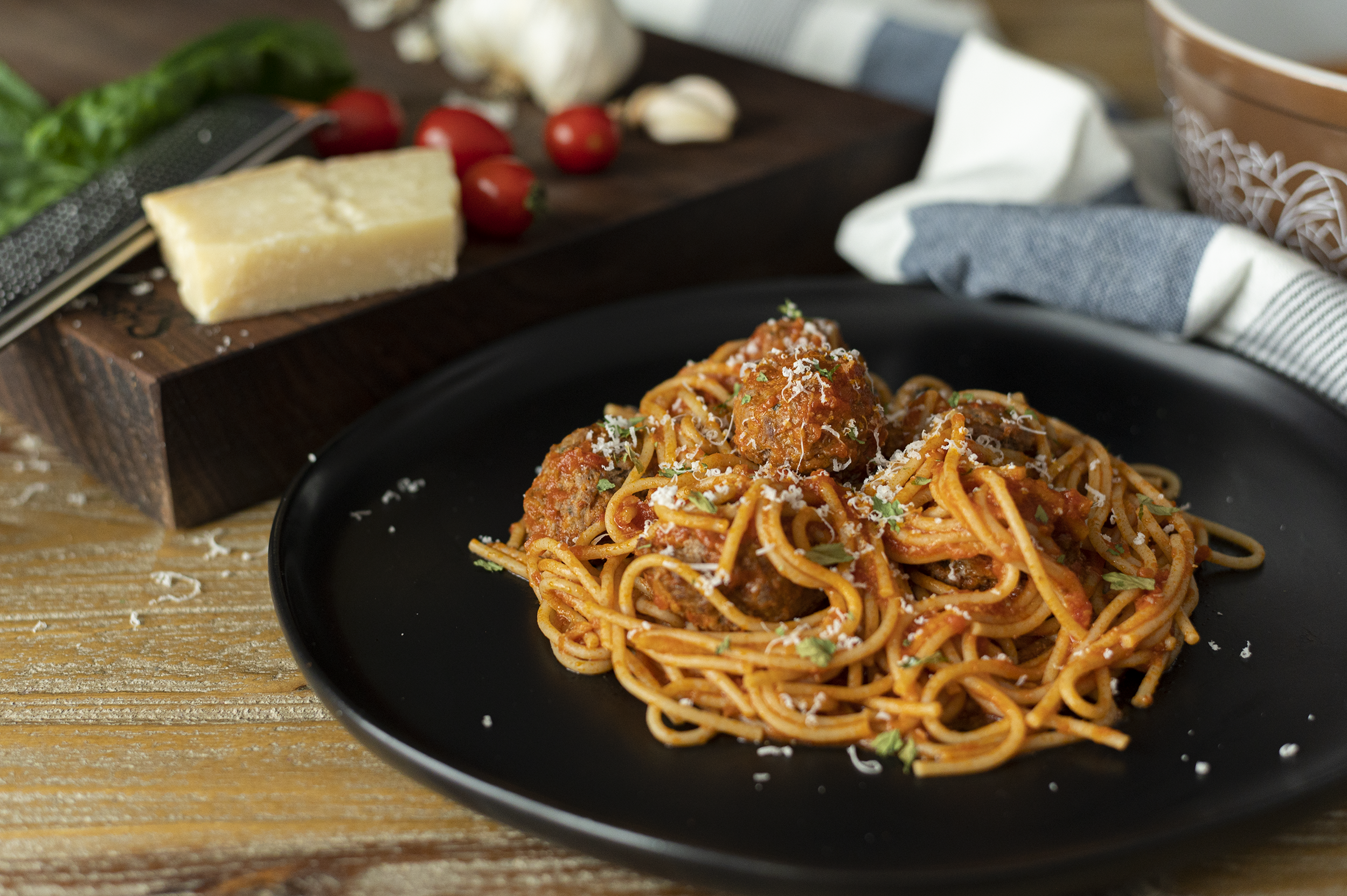 spaghetti and meatballs on a black plate on a wood table with cheese and vegetables in the background