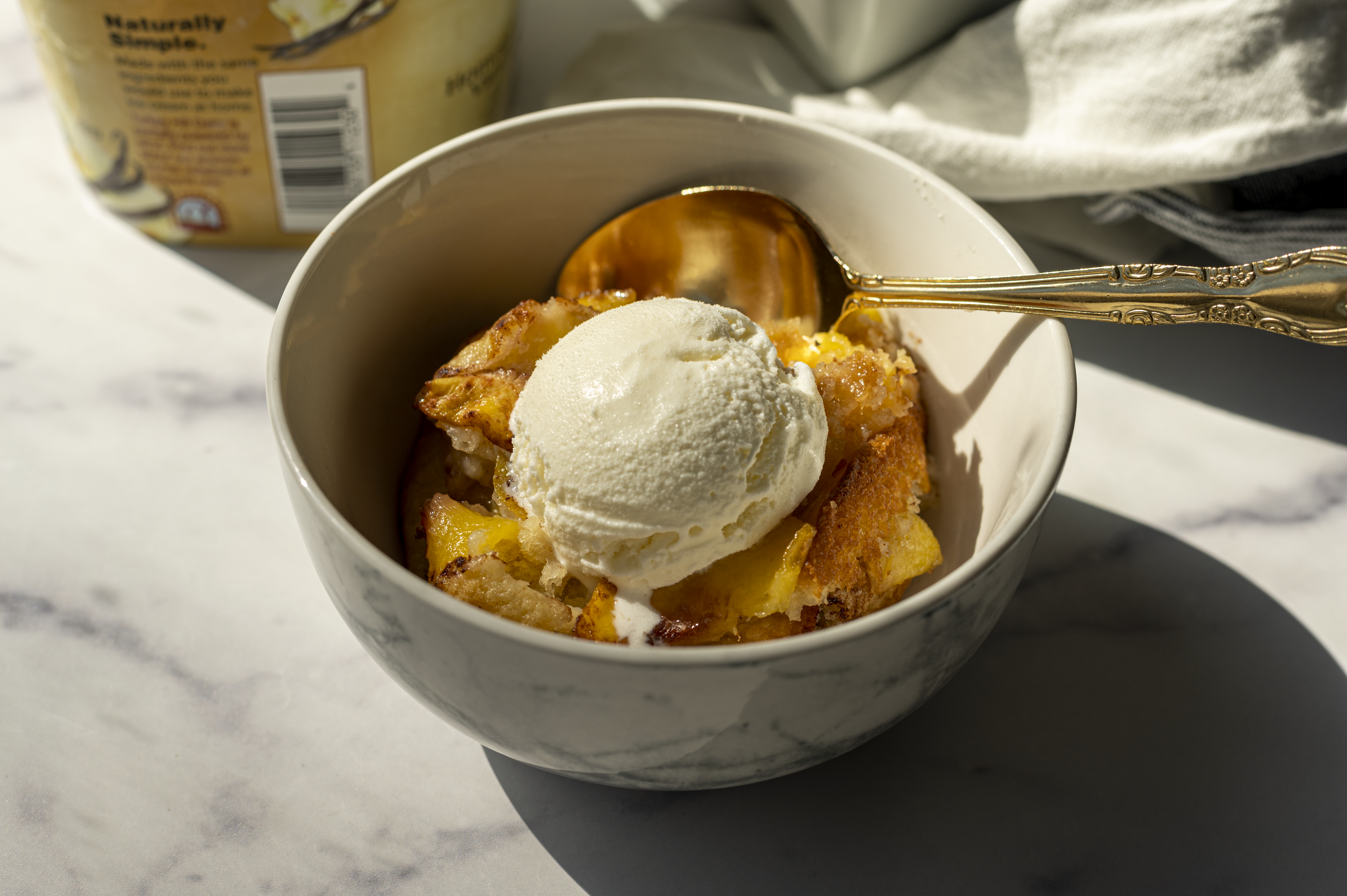 peach cobbler topped with vanilla ice cream in a white bowl against a marble background